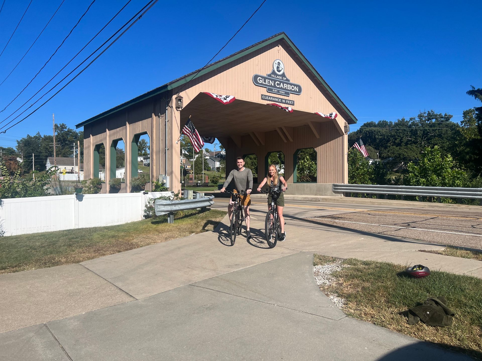 Two people biking towards a covered bridge on a sunny day.