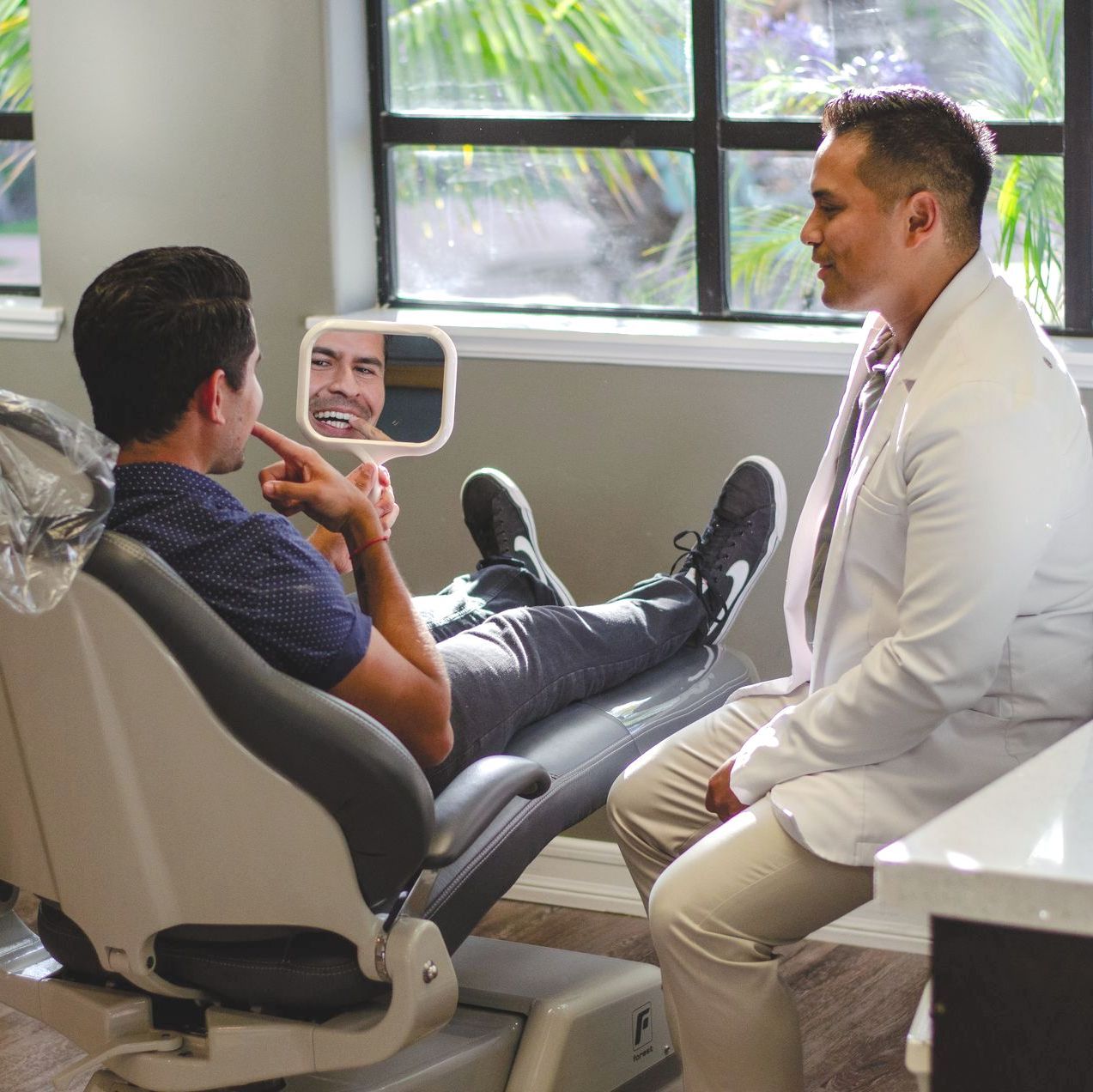 A man is sitting in a dental chair looking at his teeth in a mirror.