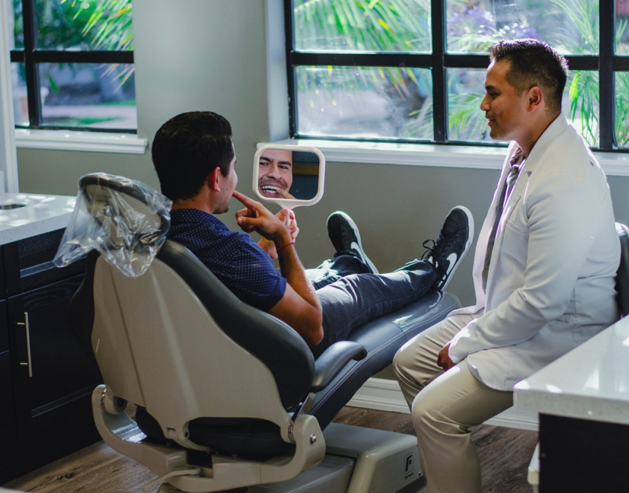 A man is sitting in a dental chair looking at his teeth in a mirror.