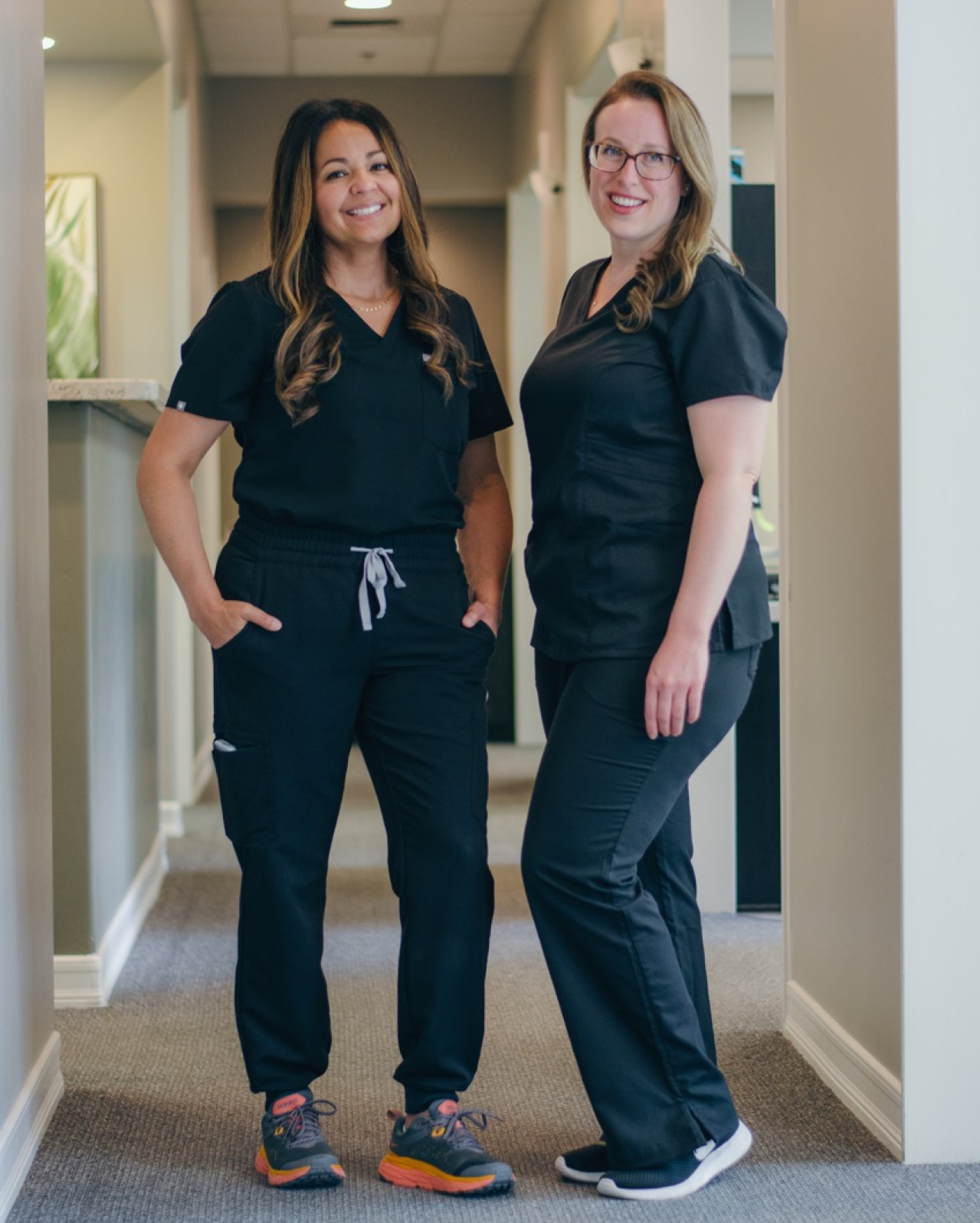 Two women in scrubs are standing next to each other in a hallway.
