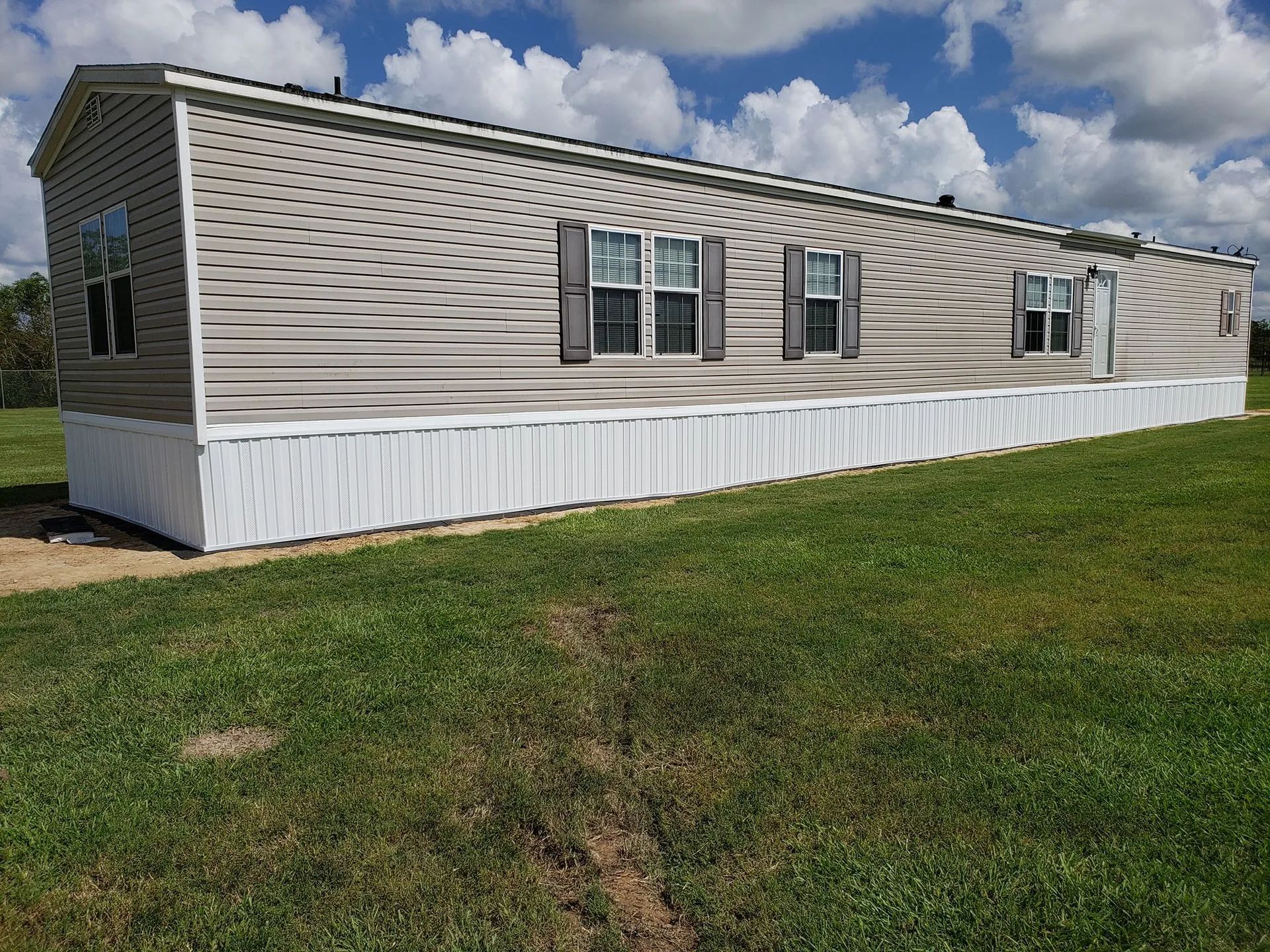 Tan mobile home with white skirting on green grass under a cloudy sky.