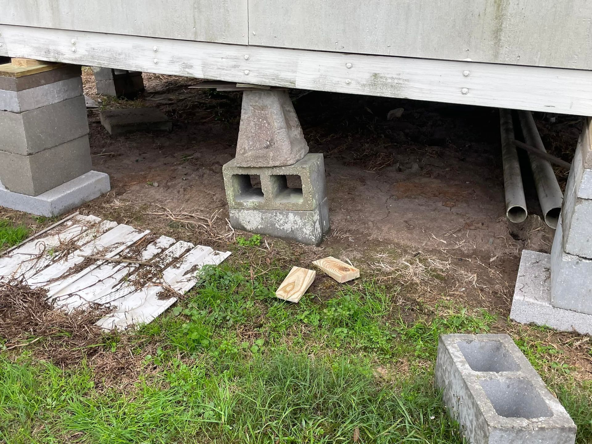 Underneath a structure supported by cinder blocks; dirt ground with a few wooden blocks and pipes present.