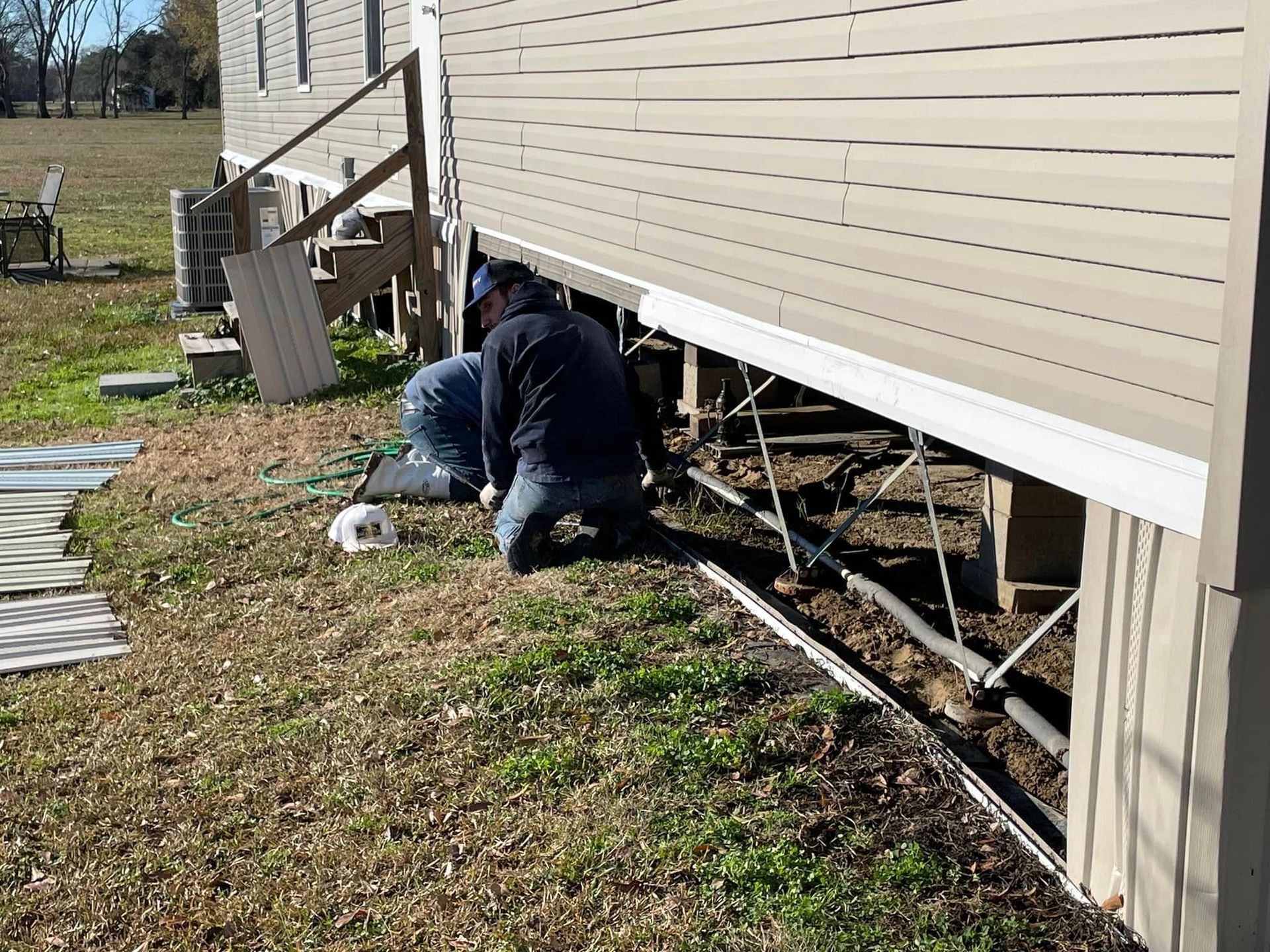 Person kneeling under a house, working on support beams. Tan siding, brown grass, clear day.