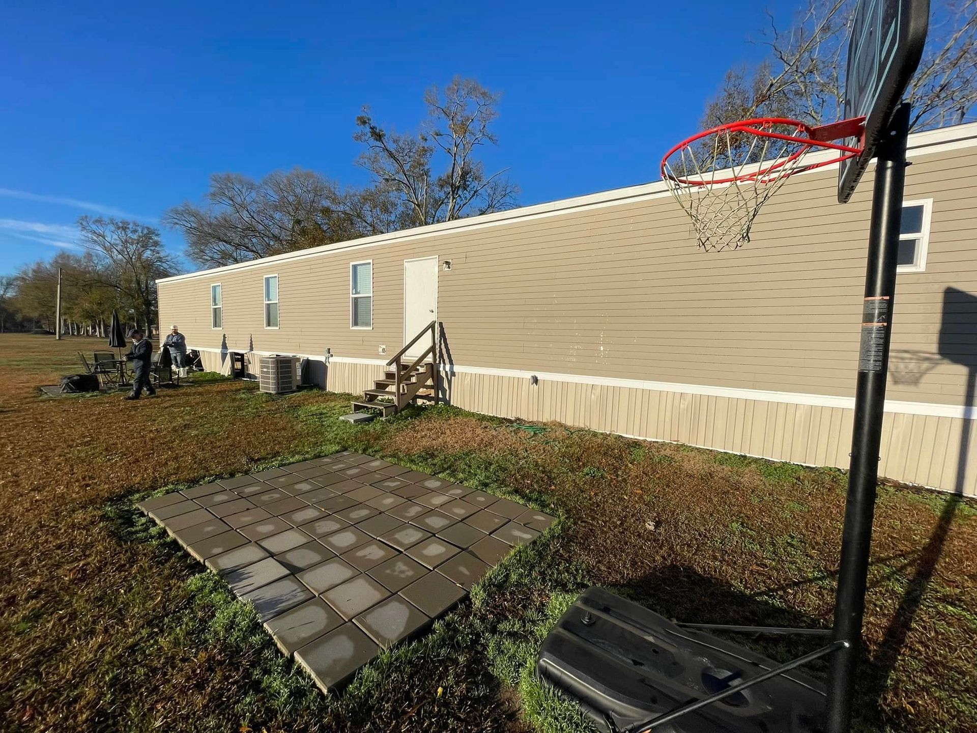 A tan mobile home with a basketball hoop, fallen panels, and people nearby on a sunny day.