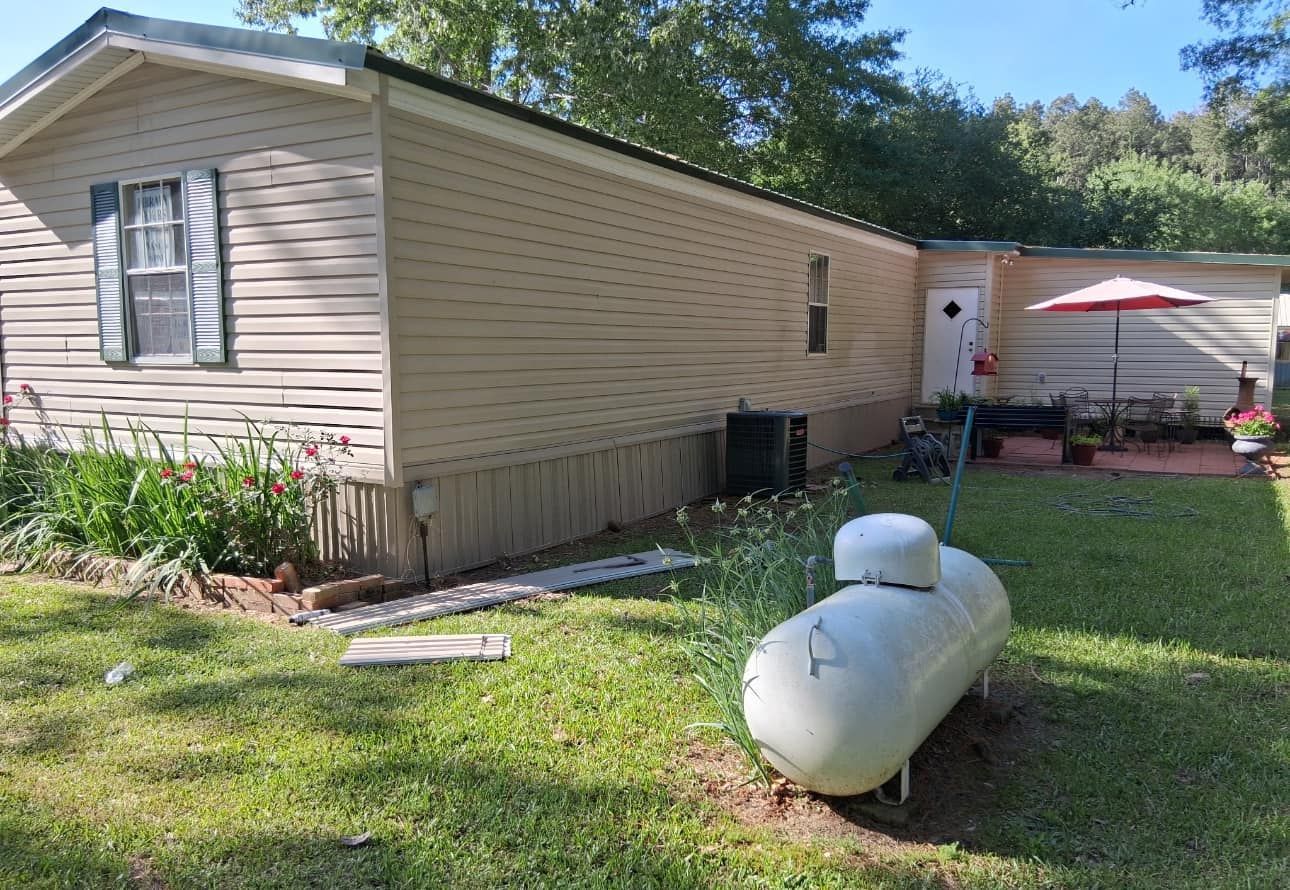 Mobile home exterior with propane tank in front yard. Green grass, beige siding, small patio, and trees.