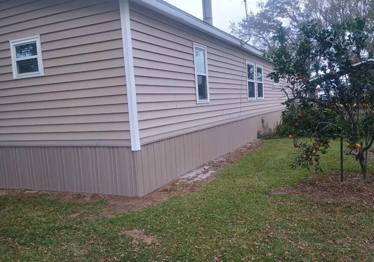 Tan siding on a mobile home with brown skirting, white trim, and a small yard.