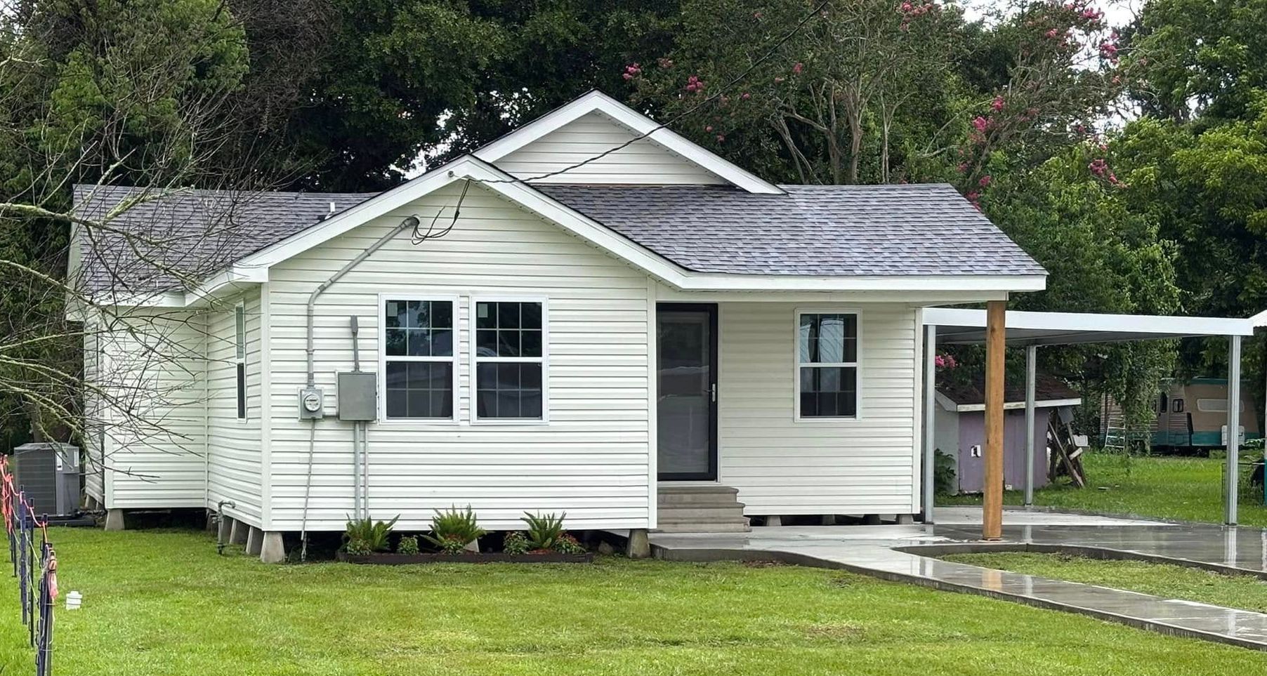 White house with a dark gray roof, small porch, and carport. The house is on a grassy lawn.