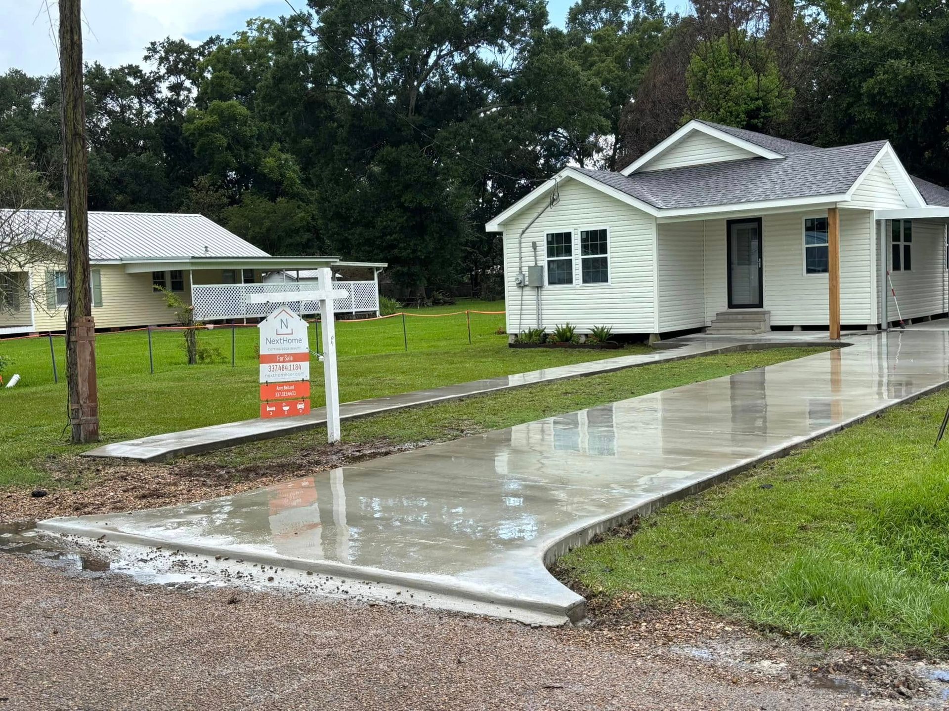 White house with a concrete driveway, a for-sale sign, and a similar yellow house in the background.