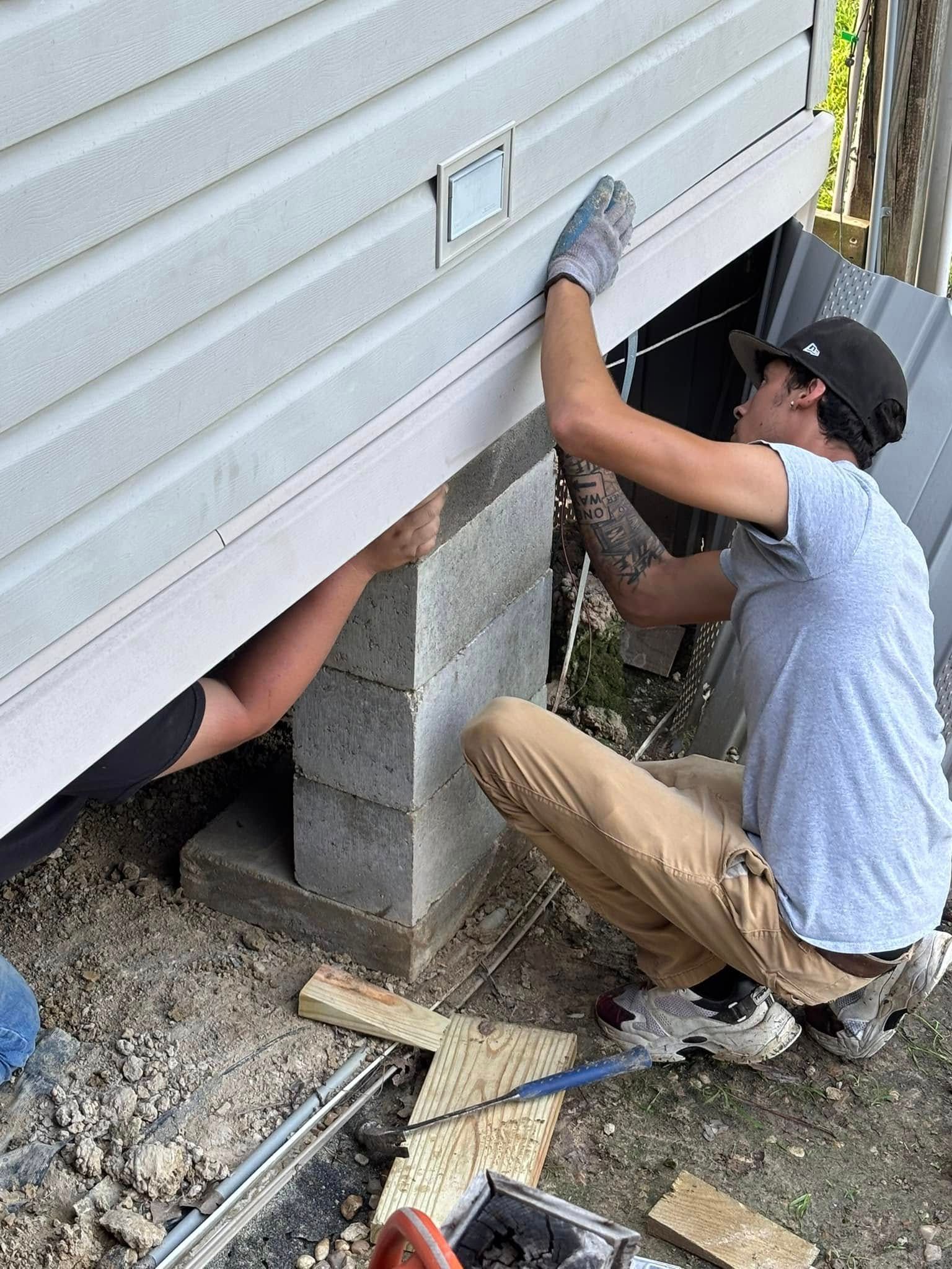 A man is kneeling down under a house while another man works on it.