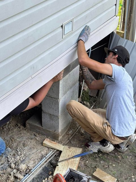 A man is kneeling down under a house while another man works on it.