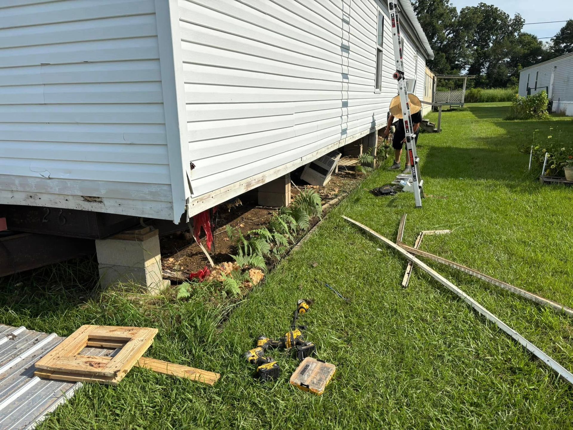 Mobile home with exposed foundation, tools and debris in a grassy yard.