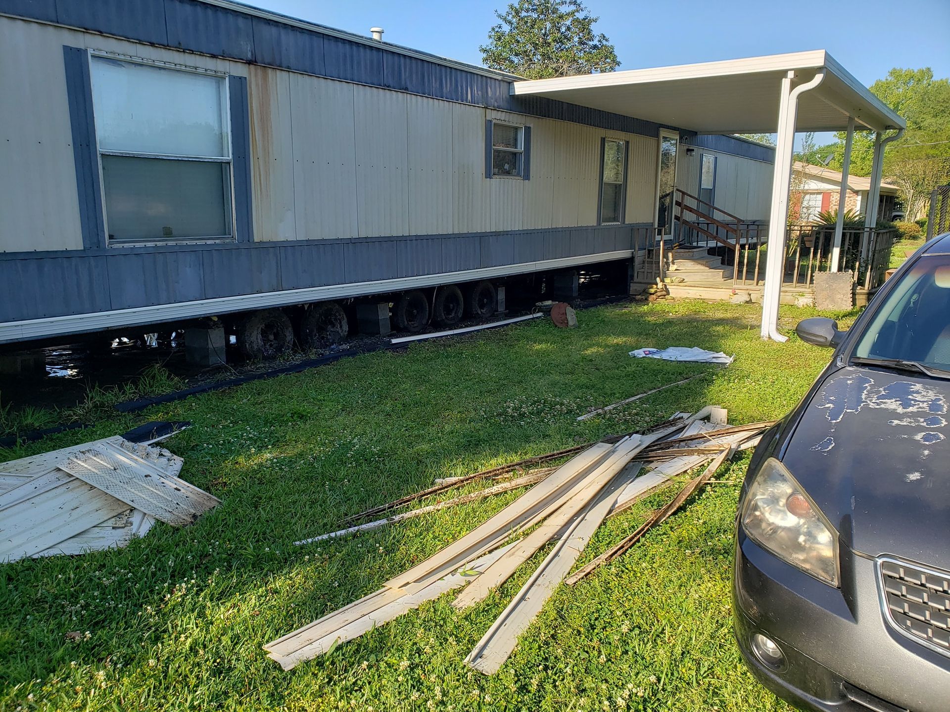 Mobile home with blue and tan siding, car parked nearby, debris on lawn.