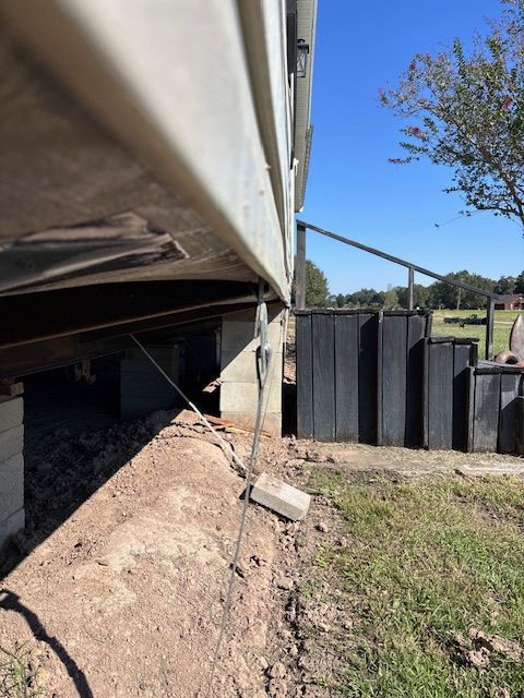 Side view of a mobile home with uneven ground and a metal support cable. Concrete blocks and stairs visible.