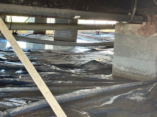 Muddy crawl space under a building, with concrete blocks supporting beams, and pipes.
