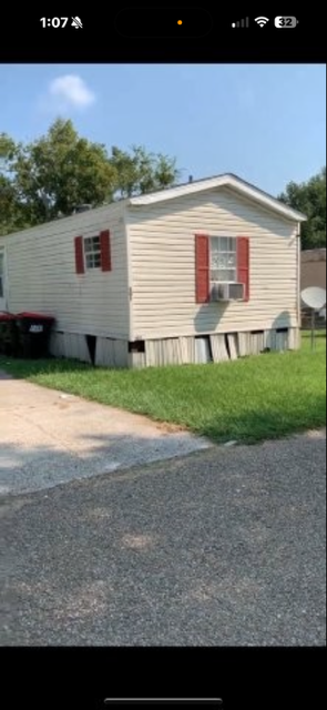 Mobile home with beige siding and red shutters, sitting on a grassy lot under a blue sky.
