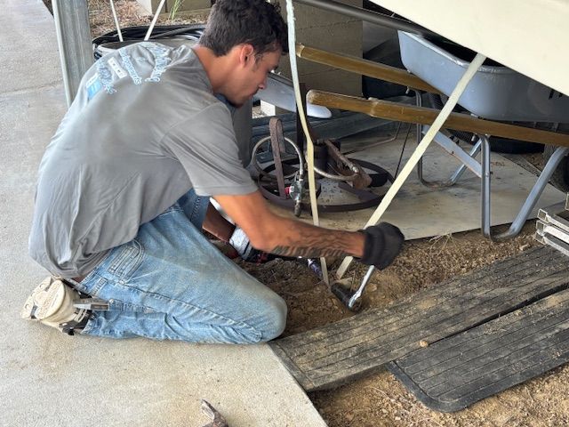 Man kneeling, working on ground, using pliers, wearing gloves. Under a carport, various tools nearby.