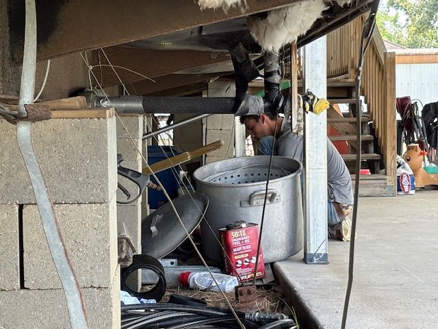 Man cooking outdoors under a mobile home, near a large pot. Gray cinder block support.