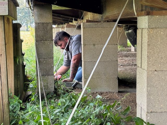 Man crouches under a building supported by concrete blocks, inspecting overgrown vegetation.