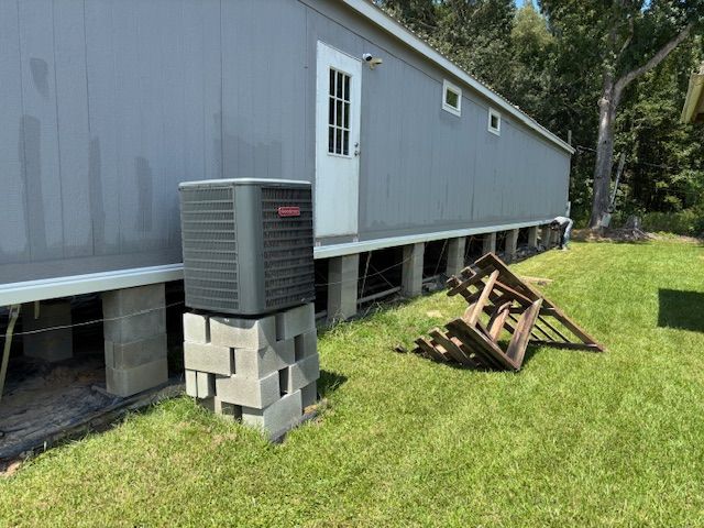 Gray mobile home with air conditioning unit on cinder blocks, door, and a wooden frame on the grass.