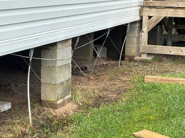 Concrete block supports under a house. Wire supports extend from the house to the ground, with grass and dirt surrounding.