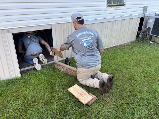 A man is kneeling down under a house while another man works on it.