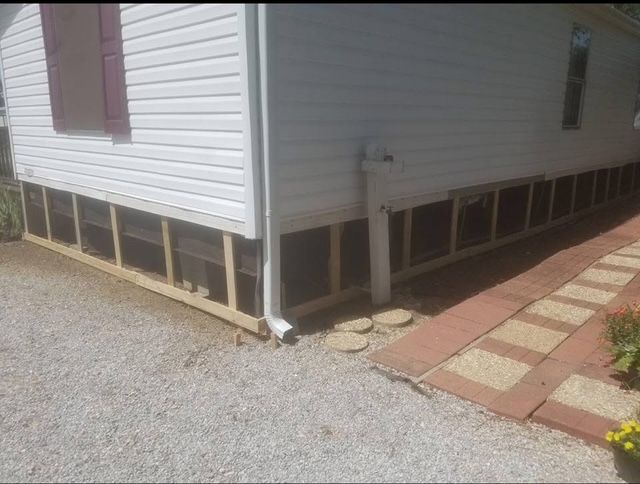 Side view of a white house with exposed wooden framing around the foundation. Gravel and brick path in front.