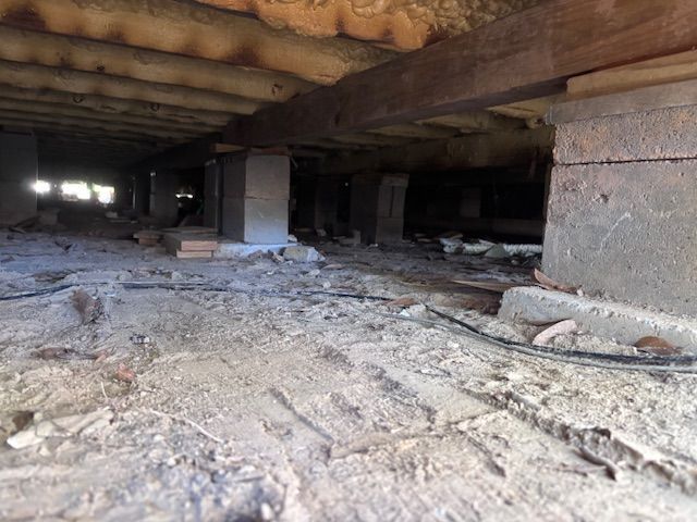 Underneath a house: dirt ground, concrete pillars support wooden beams, creating a crawl space.