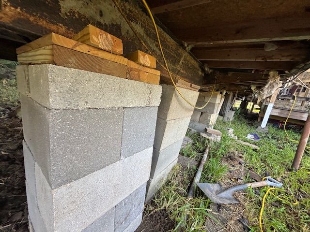 Stacked concrete blocks support a house's wooden structure. Yellow wires and a shovel are present in the grassy yard.