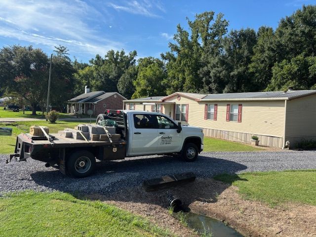 Truck parked in front of a mobile home. Gray gravel driveway, trees in the background. Bright, sunny day.