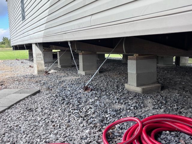 Mobile home supported by concrete blocks and anchored with cables on a gravel surface.
