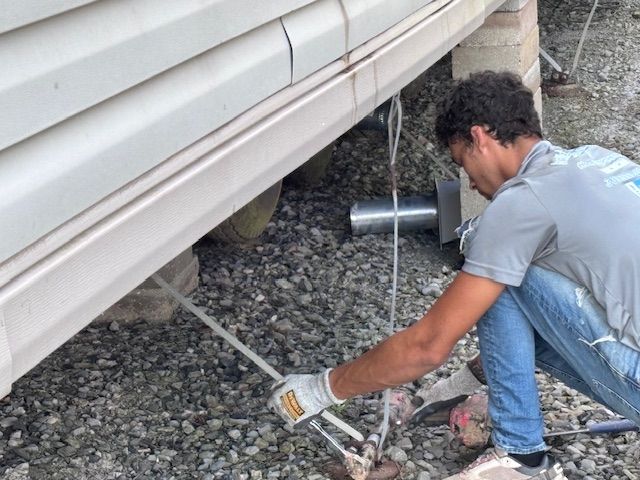 Man repairs under a house. He kneels, using tools, with gravel and a concrete base visible.