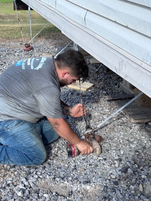Man kneeling, working under a mobile home, using a drill, gray siding, gravel ground, outdoors.