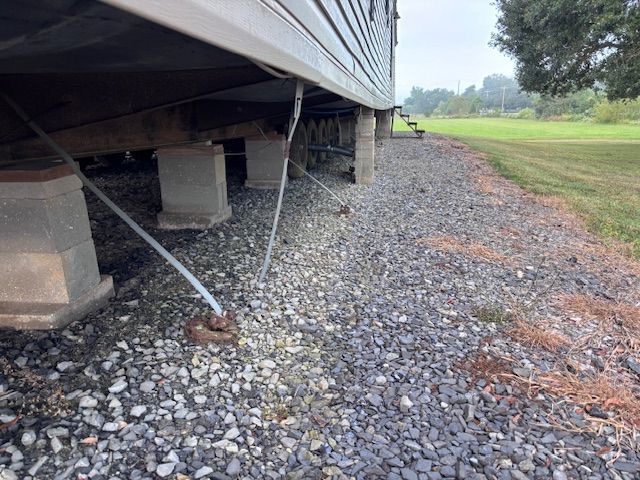 Mobile home supported by cinder blocks and guy wires on a gravel lot next to a grassy field.