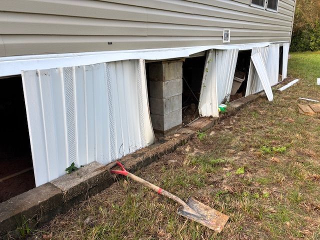 Damaged skirting around a mobile home, shovel in foreground. White metal panels are buckled and collapsed.