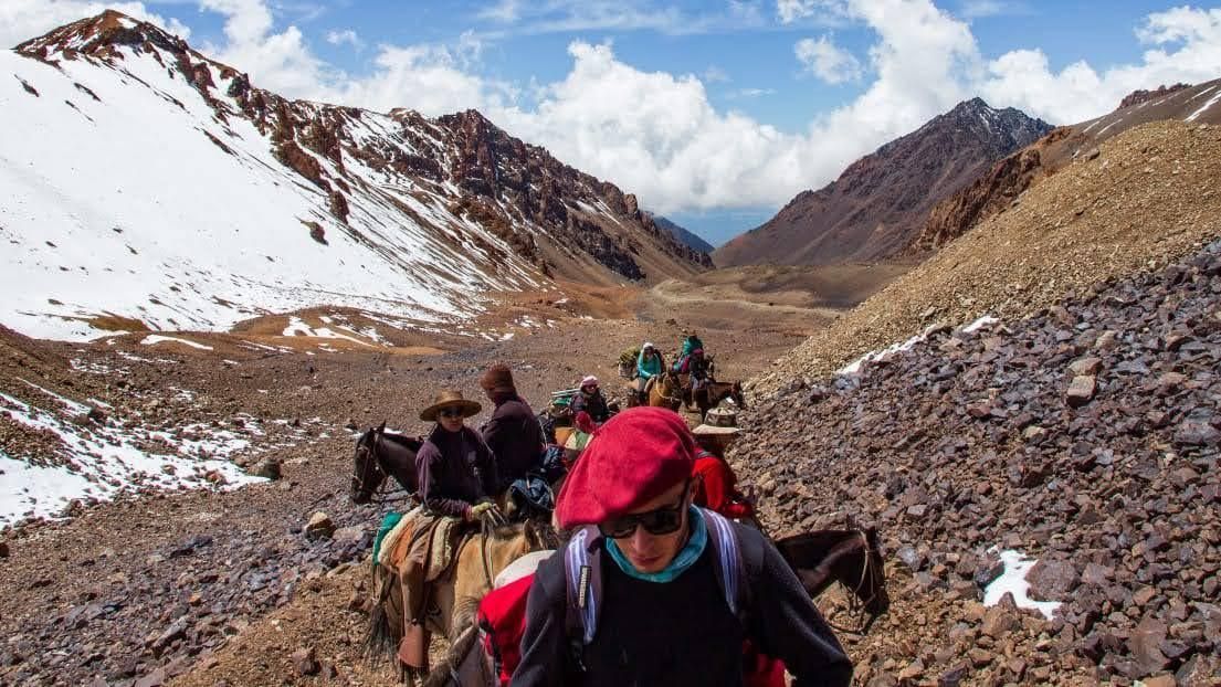 Excursionistas a caballo recorren un sendero rocoso de montaña con parches de nieve, bajo un cielo azul.