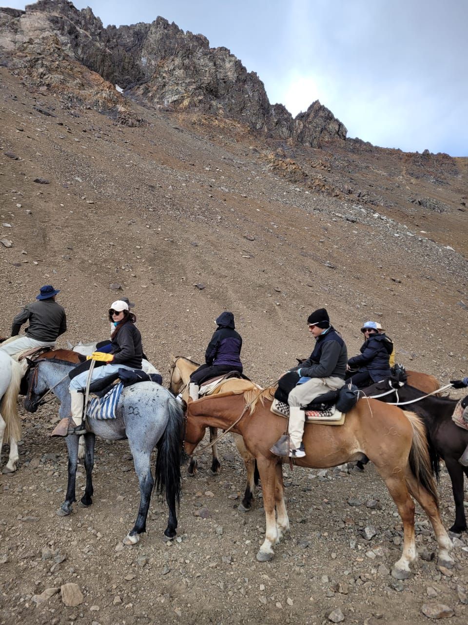 La gente a caballo se detiene en una ladera rocosa; hay montañas al fondo.