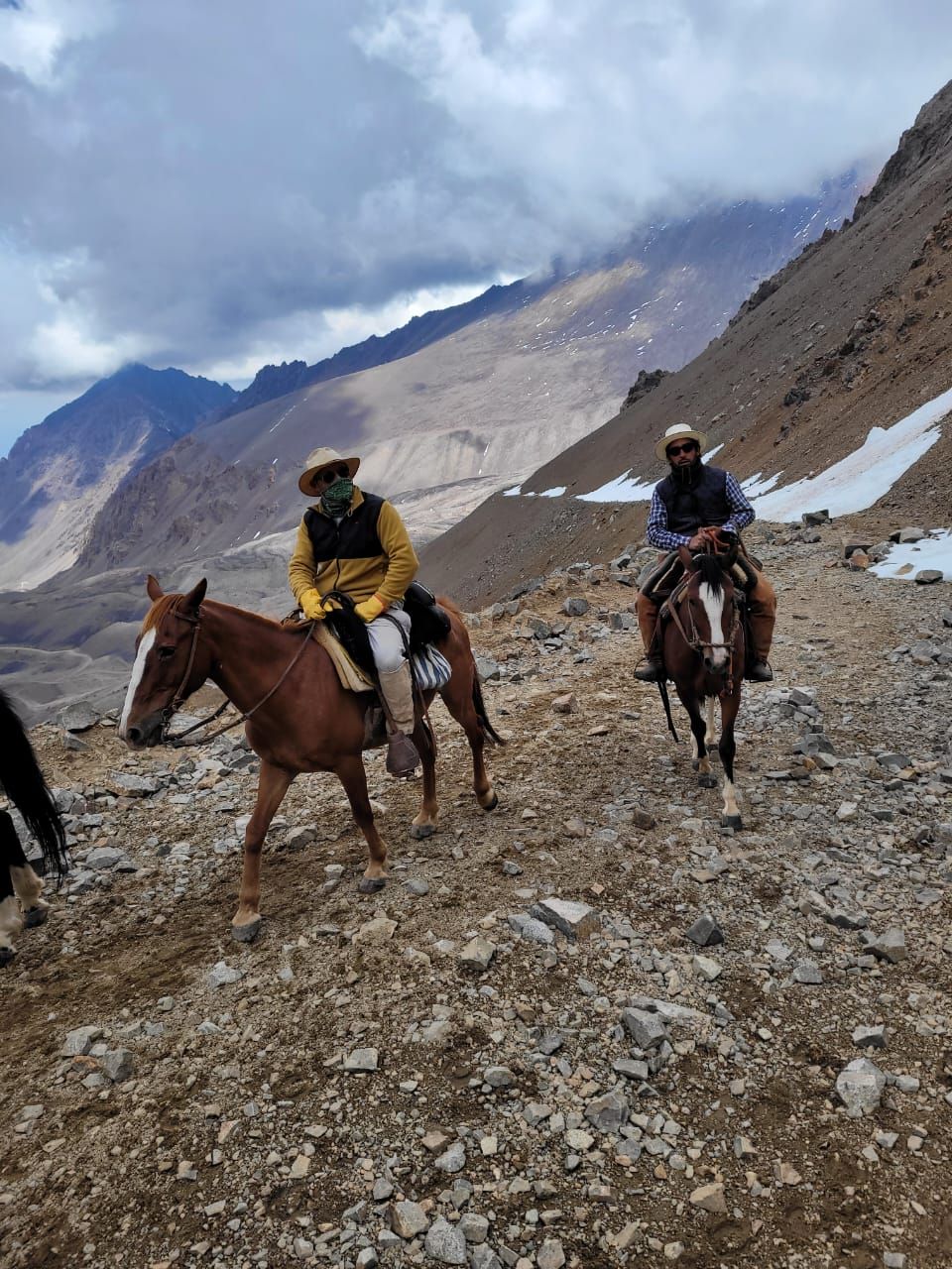Dos personas a caballo cabalgan por un sendero rocoso en la montaña, con picos nublados al fondo.