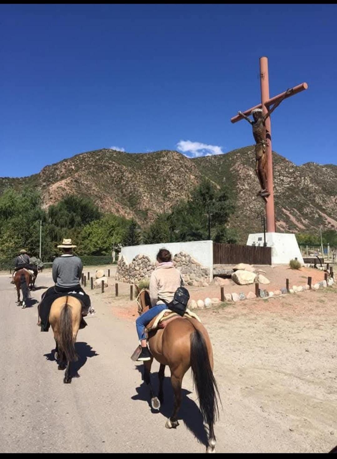 La gente a caballo pasa junto a un gran crucifijo con una montaña al fondo.