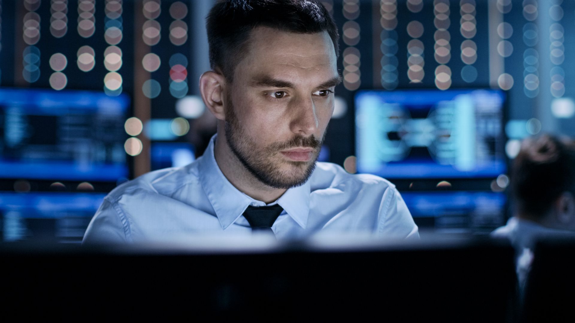 A man is sitting in front of a computer monitor in a server room.