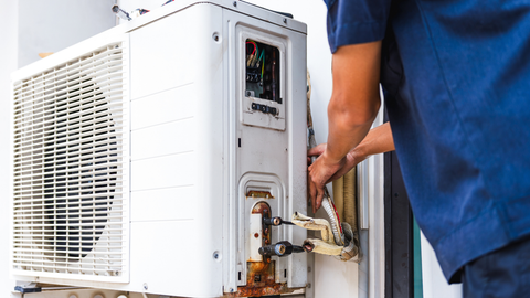 A person in blue shirt repairs an outdoor air conditioning unit with exposed wires.