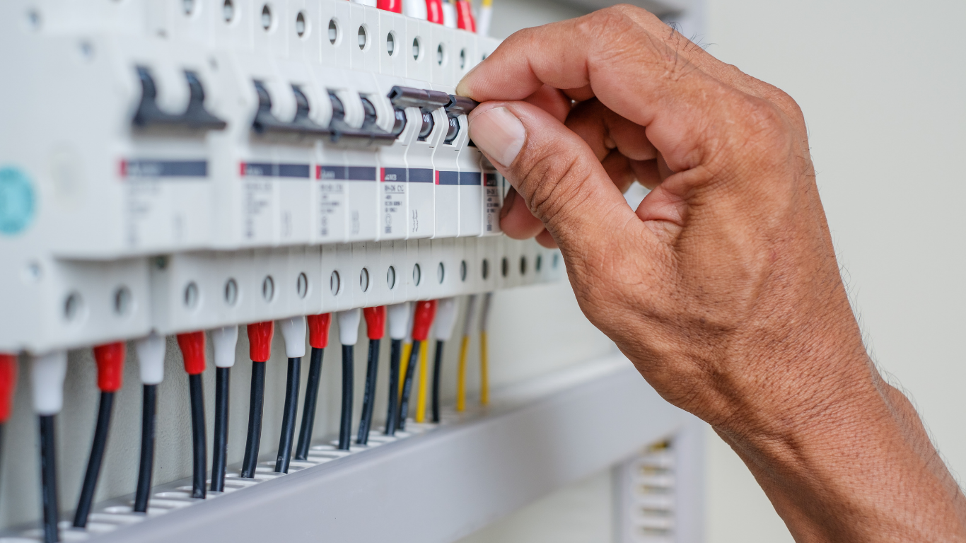 Hand flipping a circuit breaker switch in an electrical panel, with various colored wires visible.
