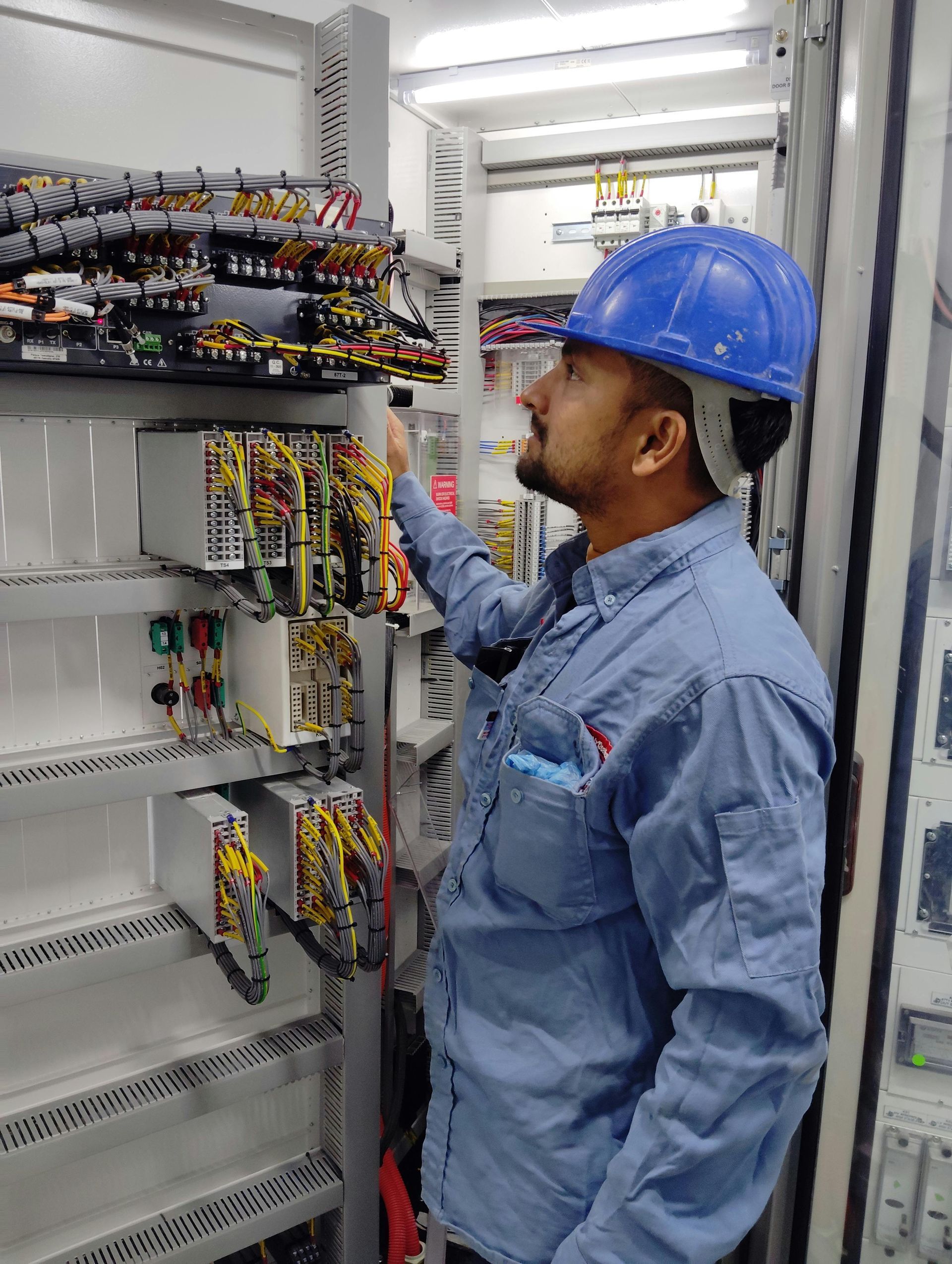 Technician in blue hard hat inspecting wiring inside an electrical control cabinet.