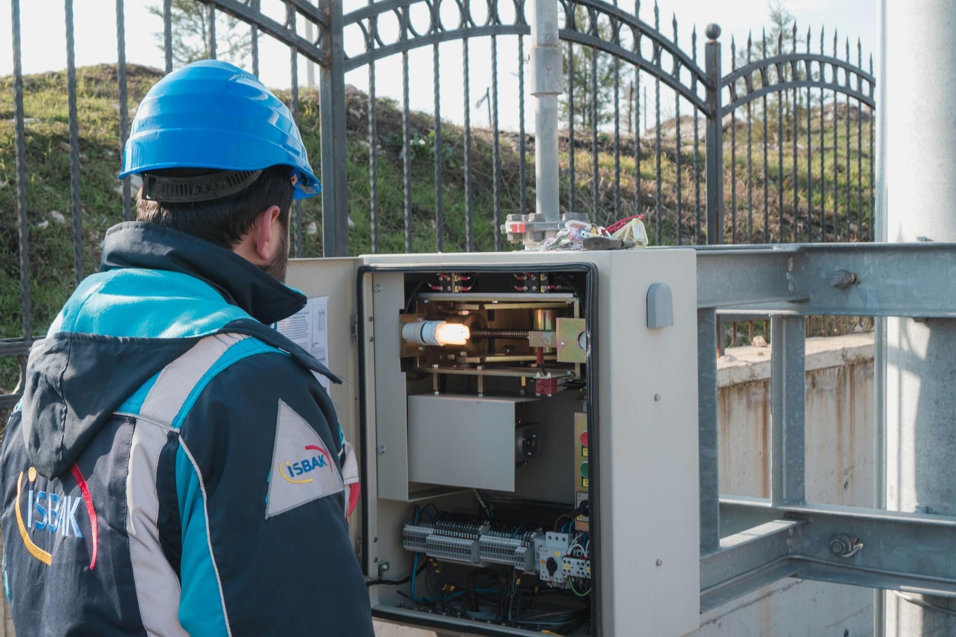 A technician wearing a blue hard hat inspects the open electrical control panel mounted on a pole outdoors.