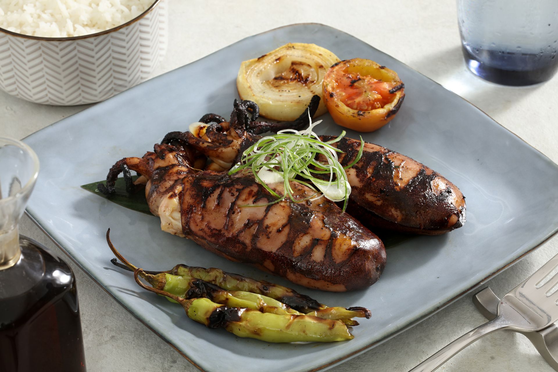Grilled squid with vegetables and herbs served on a square blue plate, accompanied by a small bowl of white rice.