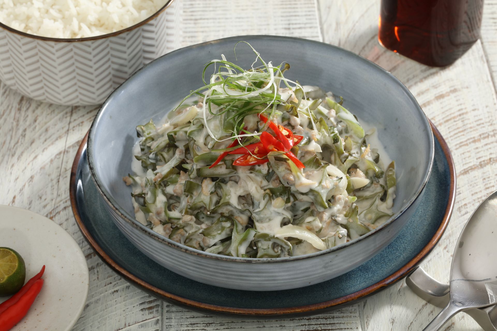A bowl of creamy Filipino bitter melon with coconut milk (ginataang ampalaya), topped with chili, beside rice and lime.
