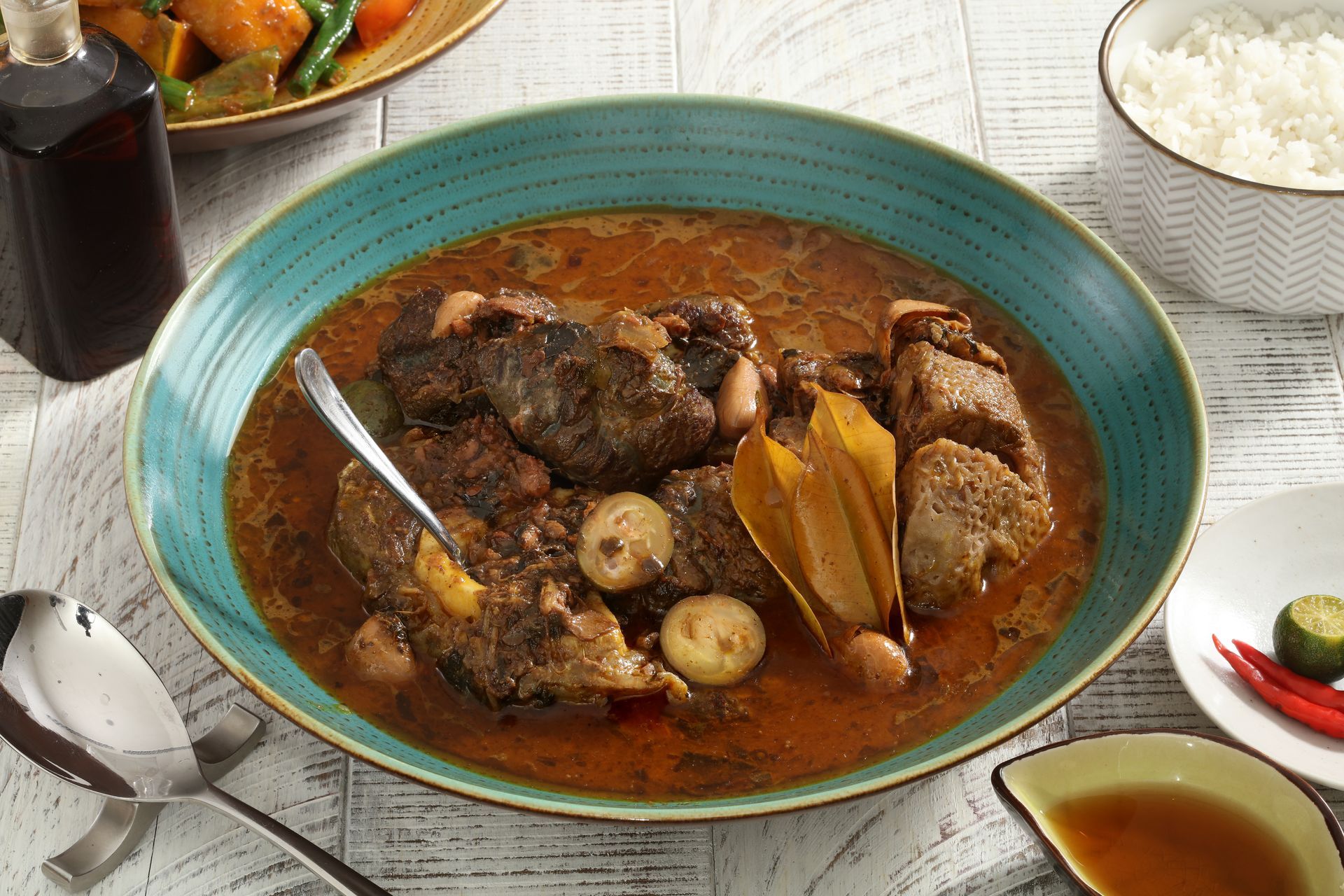 A blue bowl of Filipino beef adobo with garlic, bay leaves, and sauce, served with rice, chili, and dipping sauce.