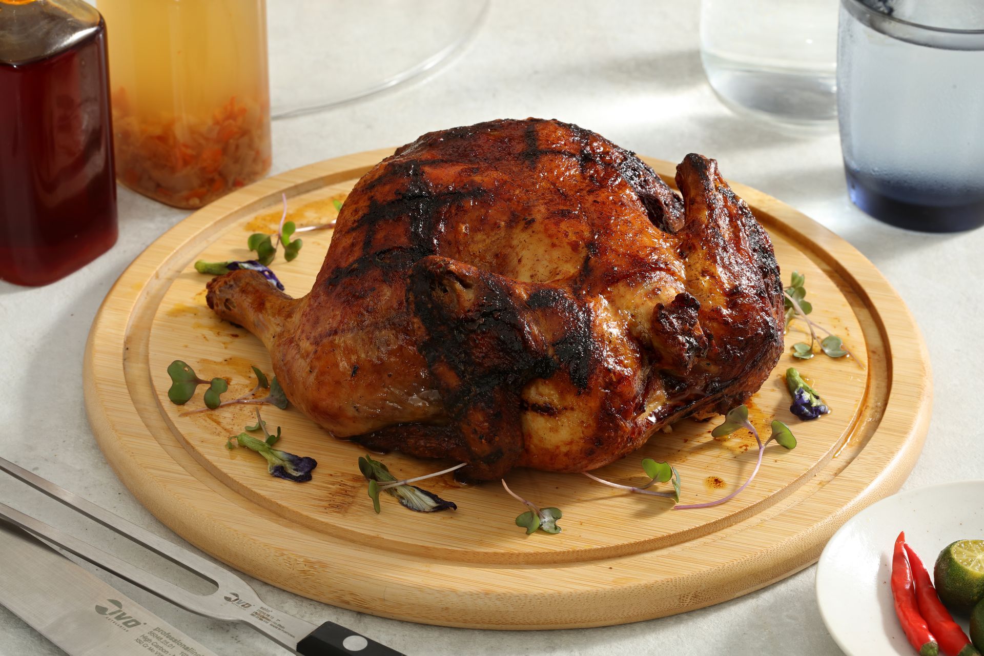 A roasted chicken with dark, glazed skin sits on a round wooden serving board with sprigs of herbs nearby.