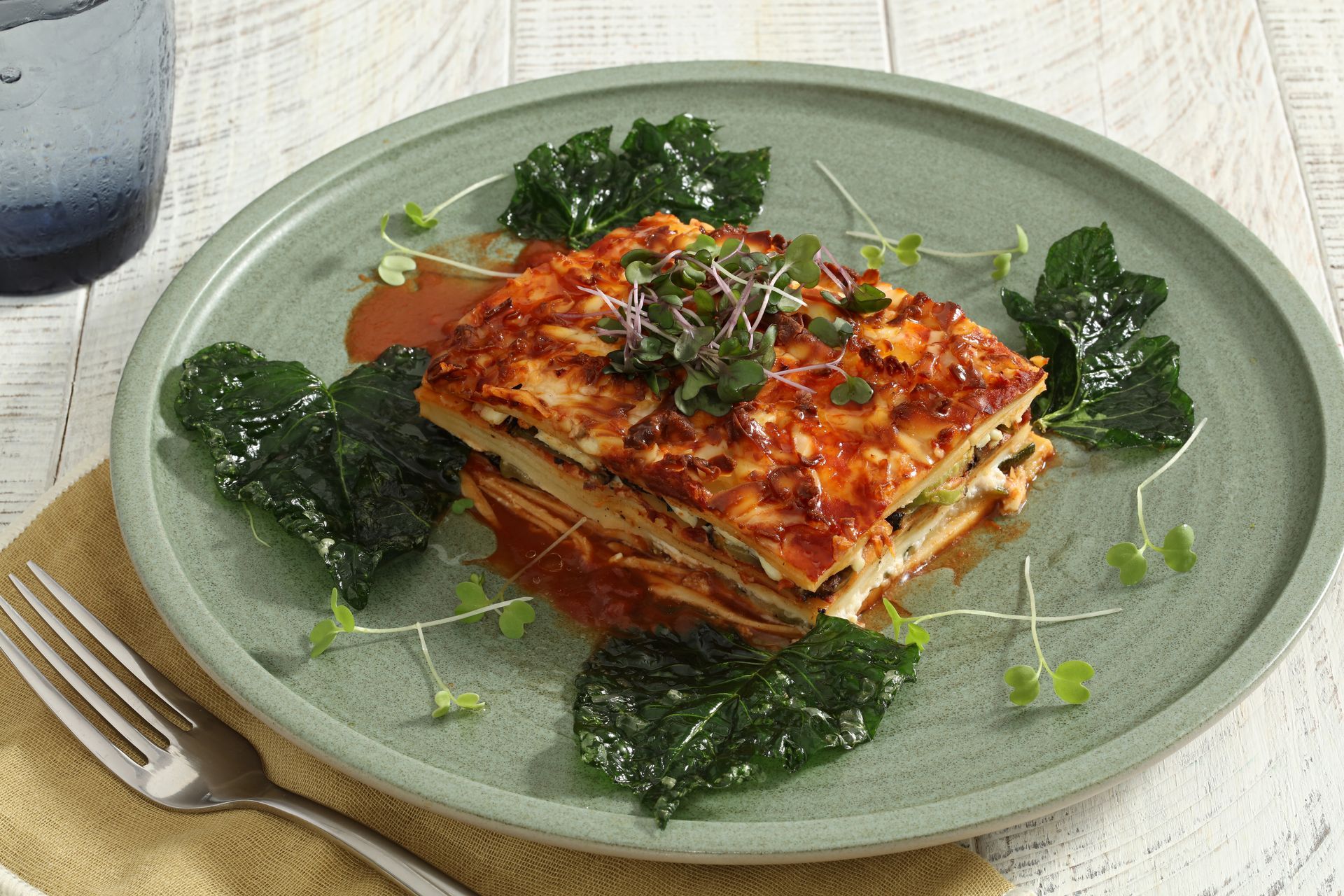 A slice of layered lasagna topped with herbs, garnished with dark leafy greens on a textured sage green plate.
