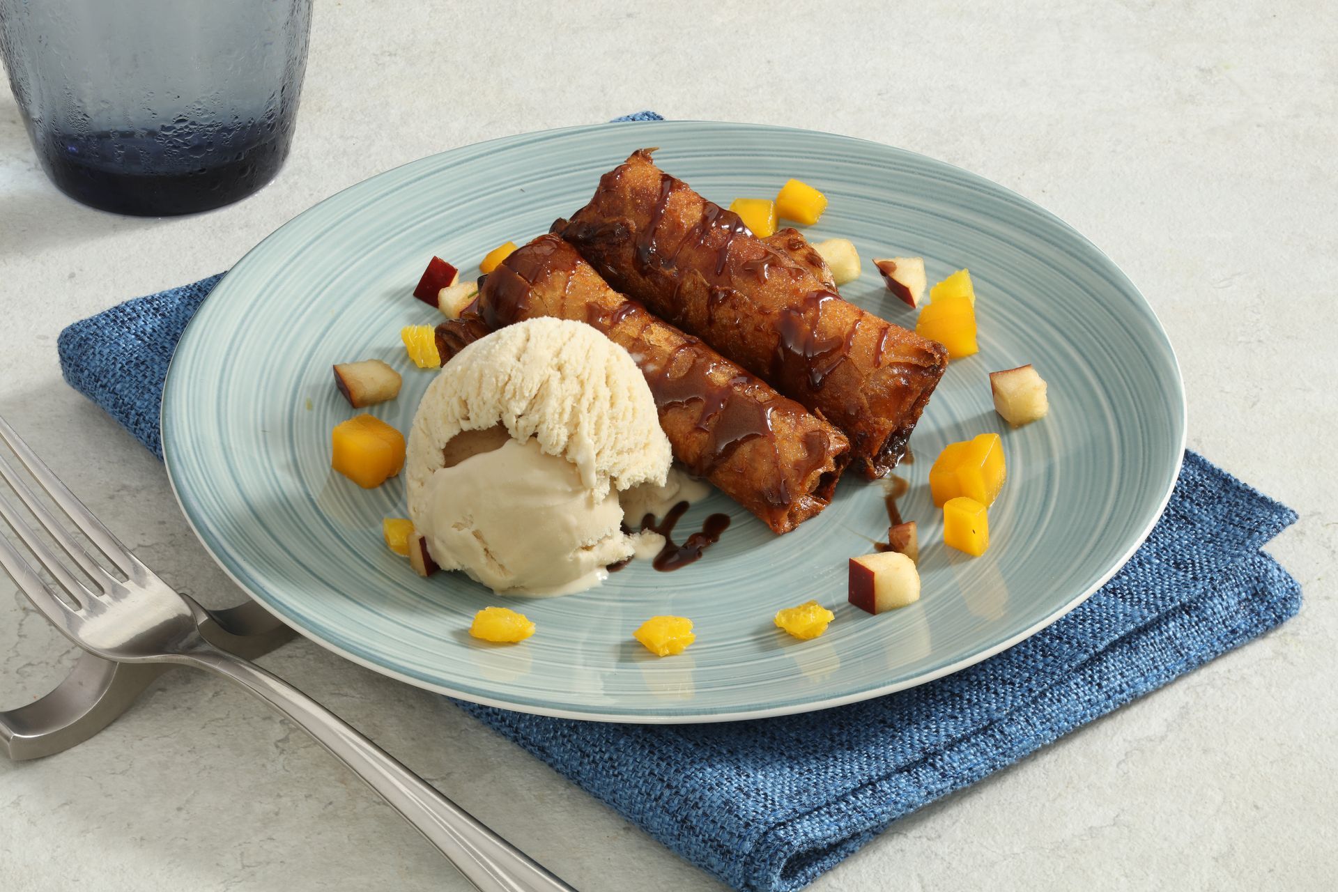 Two crispy, chocolate-drizzled dessert rolls served with a scoop of vanilla ice cream and diced fruit on a blue plate.
