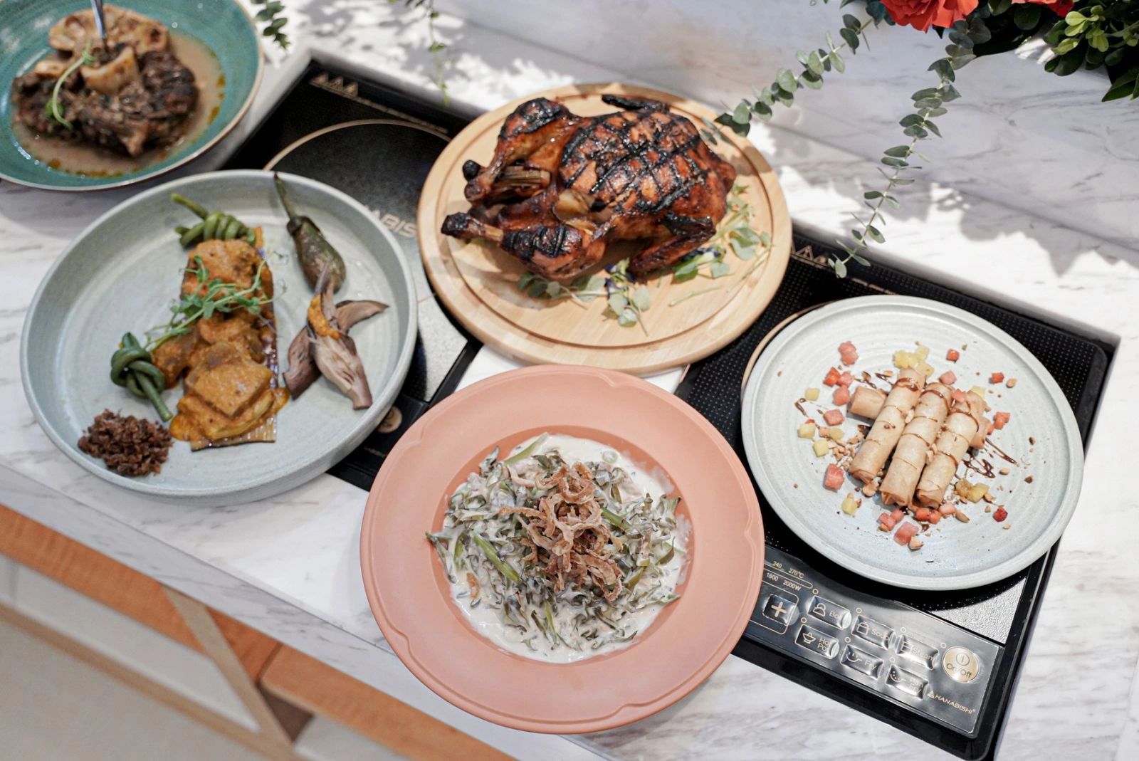 Assorted dishes on a table: steak, roasted chicken, vegetable side dishes, and spring rolls.